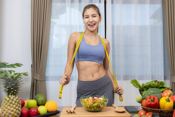 Foto colorida de mulher com fita métrica no pescoço. Ela está próximo de uma mesa com frutas, verduras e legumes - Metrópoles