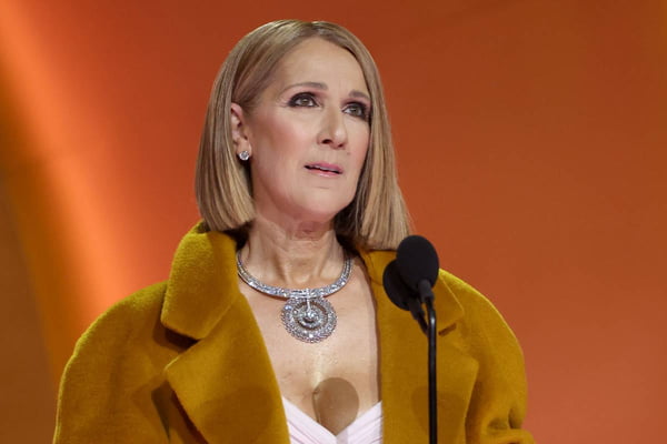 Céline Dion no Grammy 2024, com casaco longo e cabelo curto. Cantora fala em microfone - Metrópoles