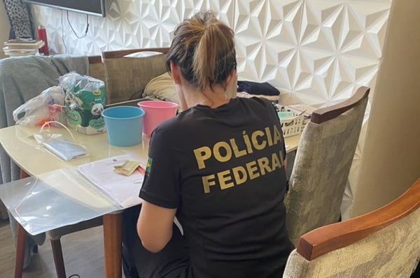 Mulher com camiseta da Polícia Federal (PF) sentada em cadeira averigua material apreendido em residência