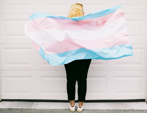 Pessoa transgênero por trás, vestindo moletom listrado rosa e branco, segura bandeira transgênero - Metrópoles