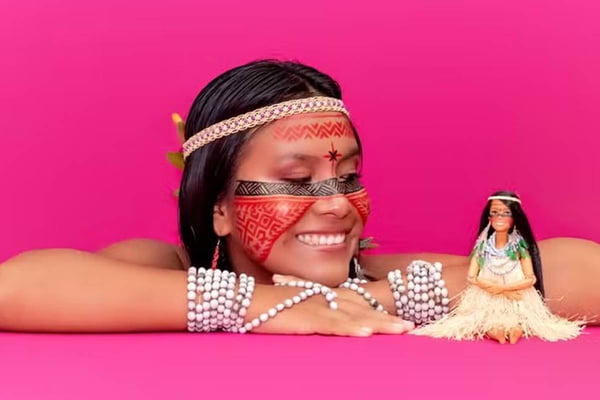 Foto colorida de uma mulher indígena ao lado de uma boneca barbie indígena em um fundo rosa - Metrópoles