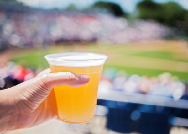 Fotografia colorida mostra mão segurando copo de cerveja e ao fundo um estádio de futebol - Metrópoles
