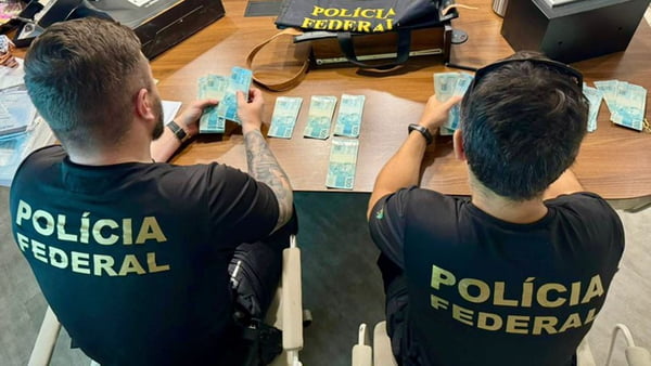 Imagem colorida das costas de dois policiais federais (PF) que estão conferindo cédulas de R$ 100 - Metrópoles