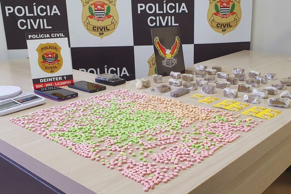 Imagem colorida de drogas apreendidas com dupla de irmãos espalhadas sobre a mesa com o banner da Polícia Civil ao fundo. Metrópoles