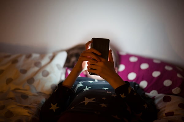 Imagem colorida de criança deitada em uma cama mexdendo no celular - Metrópoles