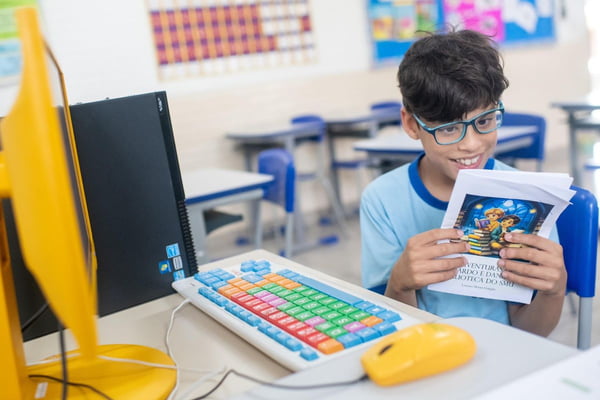 Foto colorida de criança de óculos de grau, uniforme escolar e cabelos castanhos, segurando um livro com capa colorida em frente a um computador de teclado colorido