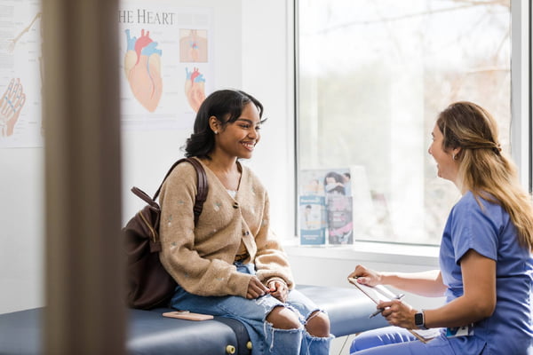 Foto colorida de paciente adulta jovem sorri enquanto ouve a enfermeira dar uma atualização encorajadora sobre seu exame médico - Metrópoles