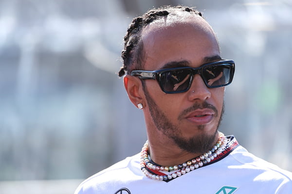 Imagem colorida de Lewis Hamilton com óculos escuros em fundo desfocado - Metrópoles