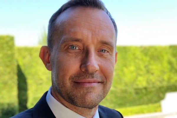 Foto colorida em close do rosto de um homem branco, loiro e de olhoso azuis sorrindo de boca fechada com um arbusto ao fundo - Metrópoles