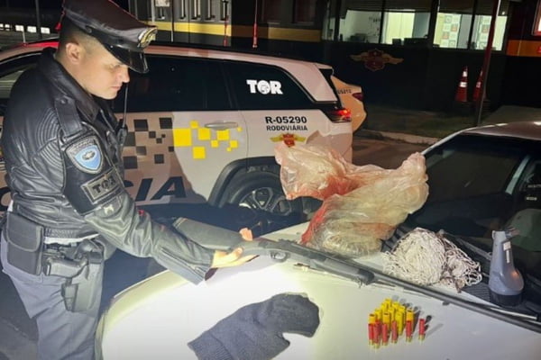 Foto colorida de policial militar diante de espingarda, munição e tatu morto sobre capô de carro - Metrópoles