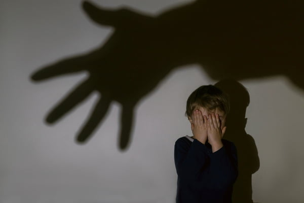 Foto colorida de garotinho e sombra de mão sobre um fundo claro. Menino cobre os olhos por medo - Metrópoles