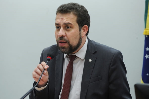 Imagem colorida mostra Guilherme Boulos, um homem pardo com cabelo e barba preto, vestindo terno cinza escuro, camisa cinza clara e gravata vinho, falando ao microfone - Metrópoles