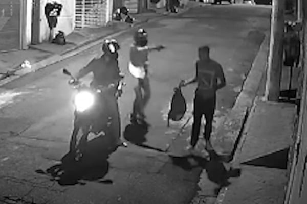 Imagem preto e branco de garupa desembarcado de moto apontando arma de fogo para rapaz que segura mochila em uma das mãos - Metrópoles