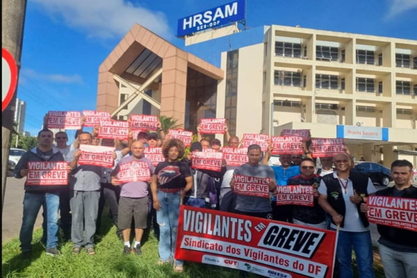 Imagem de multidão de pessoas em frente a prédio com cartazes vermelhos anunciando greve. Na placa do prédio: HRSAM