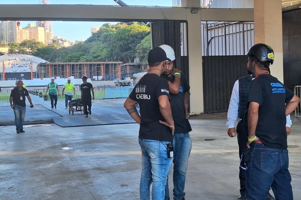 Homens com capacete de segurança conversam em frente à entrada de estádio do Pacaembu; ao fundo, homem puxa carrinho de obras