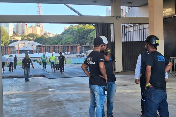 Homens com capacete de segurança conversam em frente à entrada de estádio do Pacaembu; ao fundo, homem puxa carrinho de obras