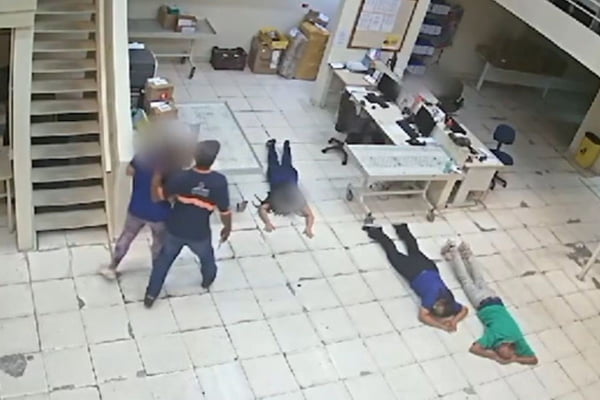 Três pessoas estão deitadas no chão de um escritório branco; um homem de boné segura outra pessoa pelo braço, ambos em pé - Metrópoles