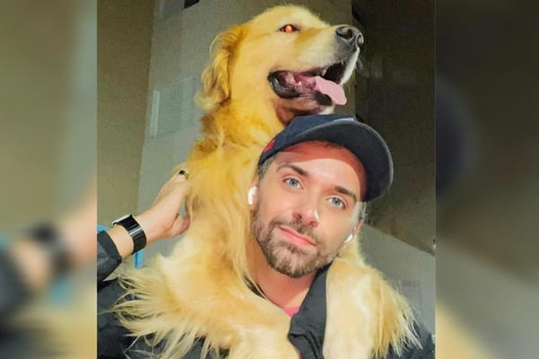 Homem branco com barba rala e boné azul segura cachorro Golden Retriever nos ombros