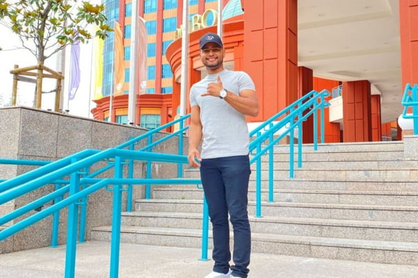 imagem colorida mostra homem jovem, com boné azul, blusa branca e calça jeans em frente a uma escada. ele faz sinal 