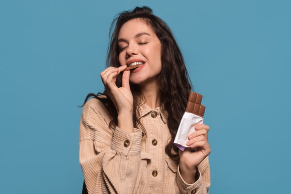 Foto colorida de mulher comendo um tablete de uma barra de chocolate. Ela segura uma barra de chocolate em outra mão - Metrópoles