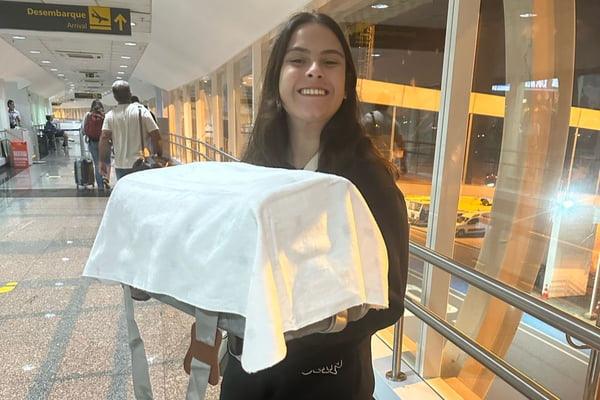 Mulher jovem branca com cabelos escuros lisos, segura com as duas mãos gaiola coberta com lençol branco. Ela está em corredor de aeroporto