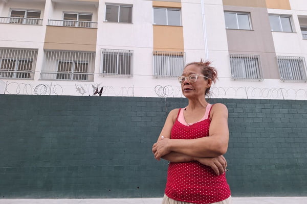 imagem colorida mostra mulher em frente a prédio de conjunto habitacional. ela tem os olhos marejados - metrópoles