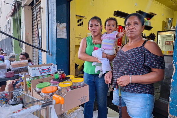 imagem colorida mostra duas mulheres e uma bebê. elas estão em frente a um carrinho de supermercado. no local, venda de balas, café e pães - metrópoles