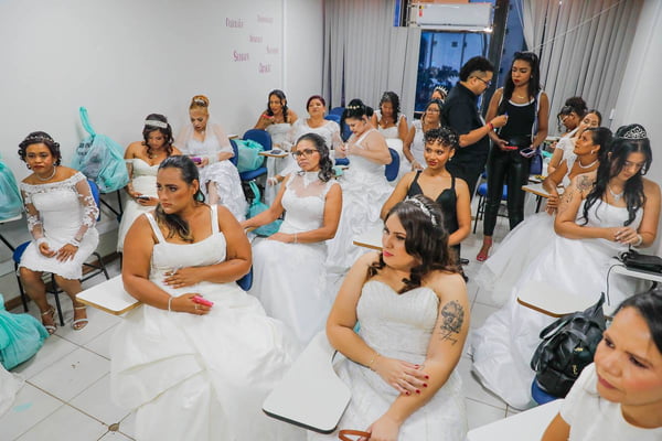 Foto colorida de 50 mulheres de vestidas de noiva sentadas em sala