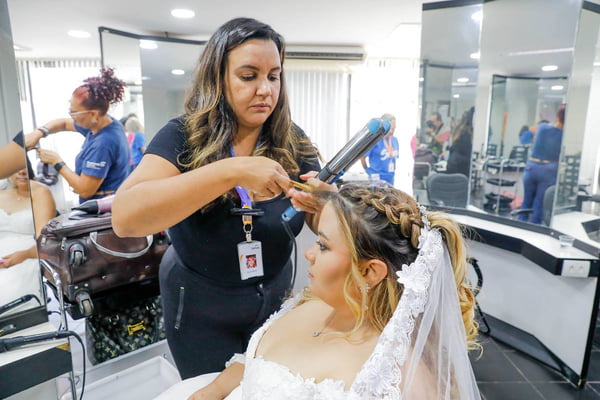 Foto colorida de cabelereira finalizando cabelo de uma mulher de vestido de noiva