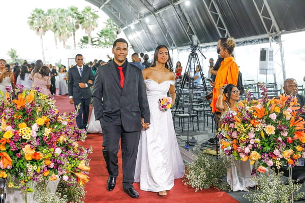 Foto colorida de casal, homem e mulher, de terno e vestido de noiva, andando em um tapete vermelho
