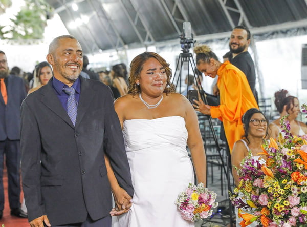 Foto colorida de casal, homem e mulher, de terno e vestido de noiva, andando em um tapete vermelho