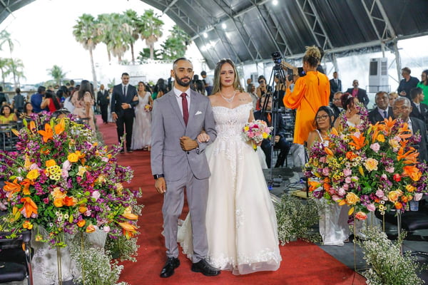 Foto colorida de casal, homem e mulher, de terno e vestido de noiva, andando em um tapete vermelho