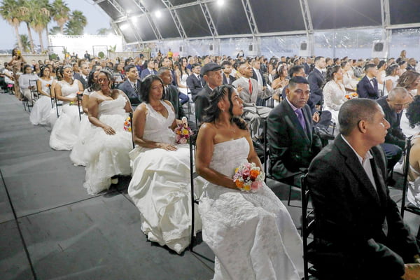 Foto colorida de plateia grande de pessoas, usando terno e vestido de noiva, sentadas