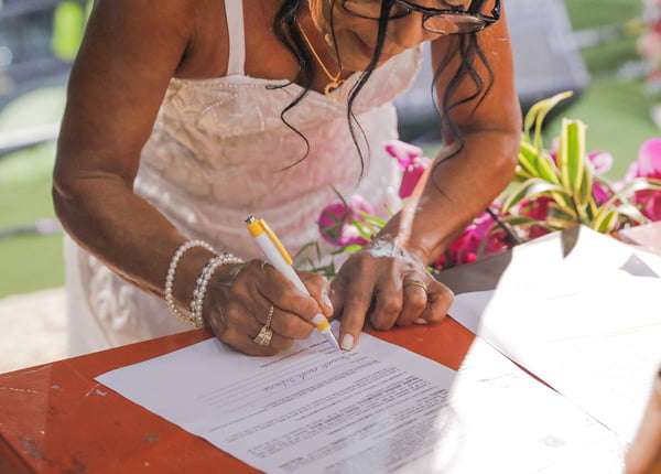Foto colorida de mulher de óculos, cabelos ondulados pretos e vestido de noiva assinando certidão de casamento