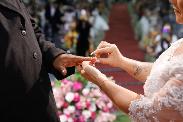 Foto colorida de casal, de casal, homem e mulher, de terno e vestido de noiva, em altar colocando a aliança no dedo do outro