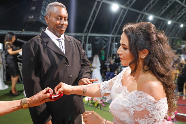Foto colorida de casal, de casal, homem e mulher, de terno e vestido de noiva, em altar, compartilhando alianças