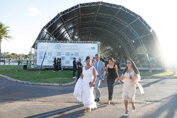 Foto colorida de estrutura de metal com toldo levantada às margens de lago. No centro da foto, uma mulher com vestido de noiva