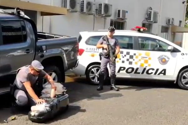 Imagem colorida de PM retirando tijolos de cocaína de tanque de combustível, ladeado por outro PM, em frente de viatura, segurando arma de fogo - Metrópoles
