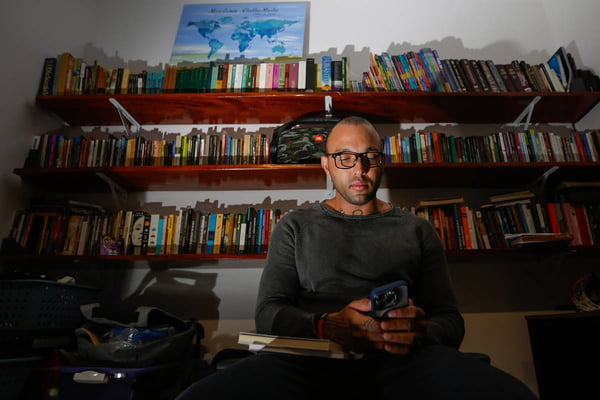 Foto colorida de homem negro em quarto com livros e mãos machucadas