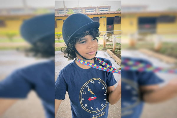 Foto colorida de criança com capacete de hipismo e cordão com símbolo do autismo