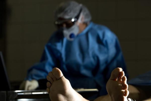 Médico legista ao fundo com o pé de cadáver em primeiro plano