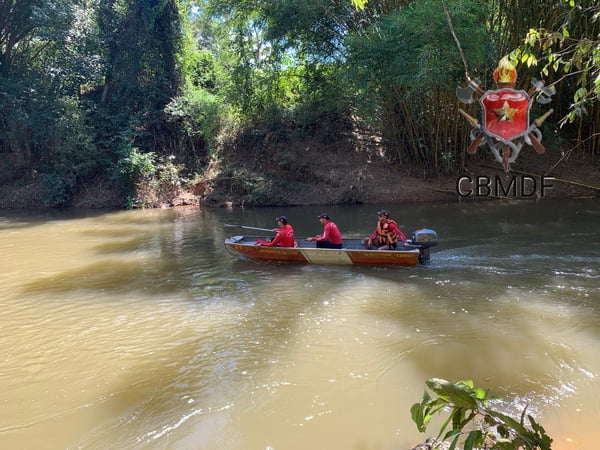 Foto colorida tirada de dia de barco navegando em meio a um rio, tripulado por três homens com uniforme de bombeiro