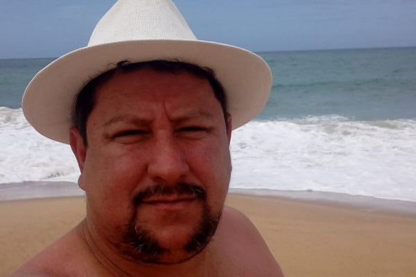 Foto colorida de homem branco, com sobrepeso e cavanhaque, usando chap&eacute;u em dia ensolarado em frente ao mar - Metr&oacute;poles