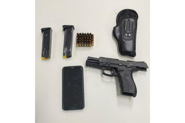 Foto colorida de pistola, dois carregadores, coldre e celular, todos pretos, sobre mesa branca - Metrópoles