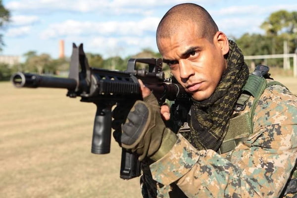 Foto de um homem negro, careca, com roupa do exército, segurando uma arma - Metrópoles