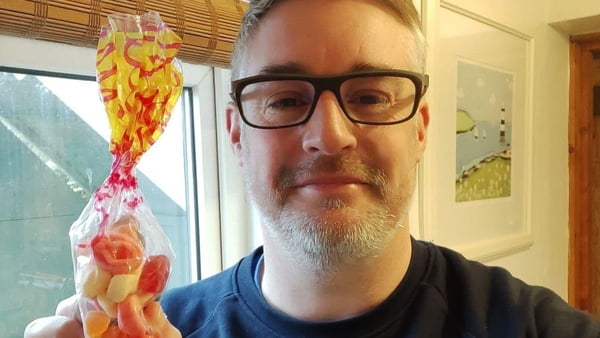 homem branco de óculos e barba segurando um saquinho de doces coloridos