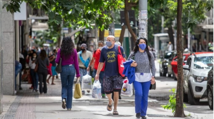Fotografia colorida. Pessoas caminham em calçada com máscaras e sacolas nas mãos