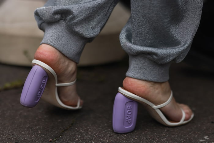 Mulher branca chegando na Semana de Moda de Paris em março de 2022. Na imagem é possível ver apenas o pé da pessoa, que usa uma sandália branca da marca Loewe, que possui um salto grosso lilás imitando um sabonete. - Metrópoles