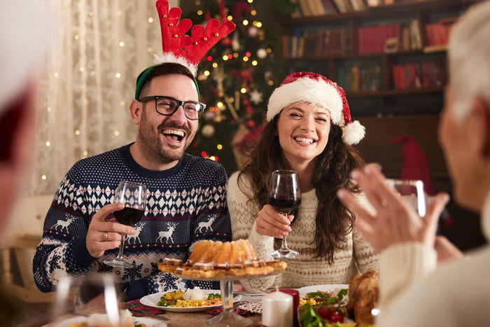 Foto colorida de homem com chapéu natalino ao lado de mulher com gorro do Papai Noel. Eles estão com taças de vinho na mão e diante de uma mesa com vários pratos - Metrópoles