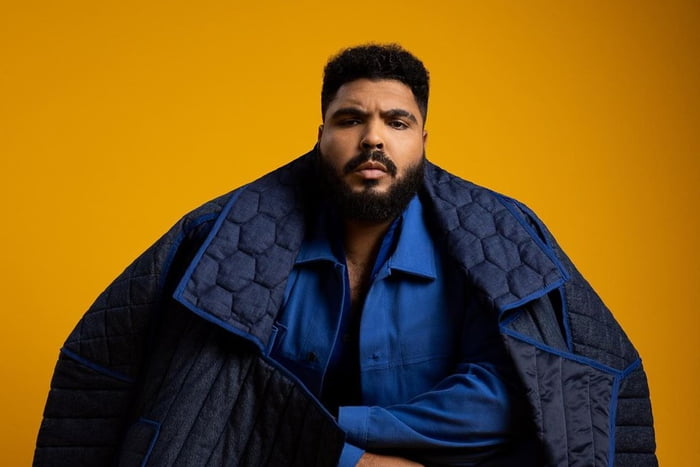 O humorista Paulo Vieira posa para foto com jaqueta estilizada, de braços cruzados sob fundo amarelo - Metrópoles